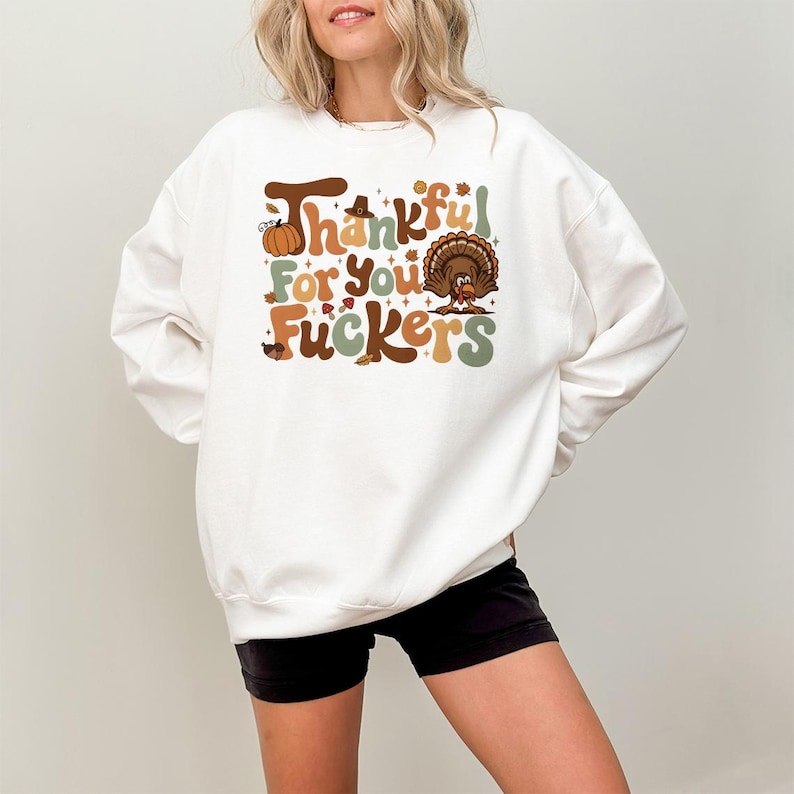 Op de afbeelding: Witte sweatshirt met een Thanksgiving-thema grafisch ontwerp. Het ontwerp bevat de tekst "Thankful For You Fuckers" in bruin, oranje en groen, samen met een pompoen en een kalkoen illustratie. Het sweatshirt is een casual kledingstuk met lange mouwen.