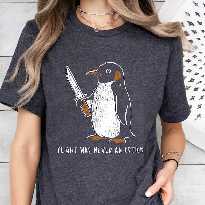 May include: Dark gray t-shirt featuring a cartoon penguin holding a knife. The penguin is black and white with orange accents. The text "FLIGHT WAS NEVER AN OPTION" is printed below the penguin.
