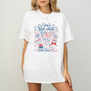May include: White t-shirt with a patriotic graphic in red and blue. The design includes an American flag, fireworks, and the text "Red, white and Due". Other illustrations include a baby bottle, pacifier, and cherries.