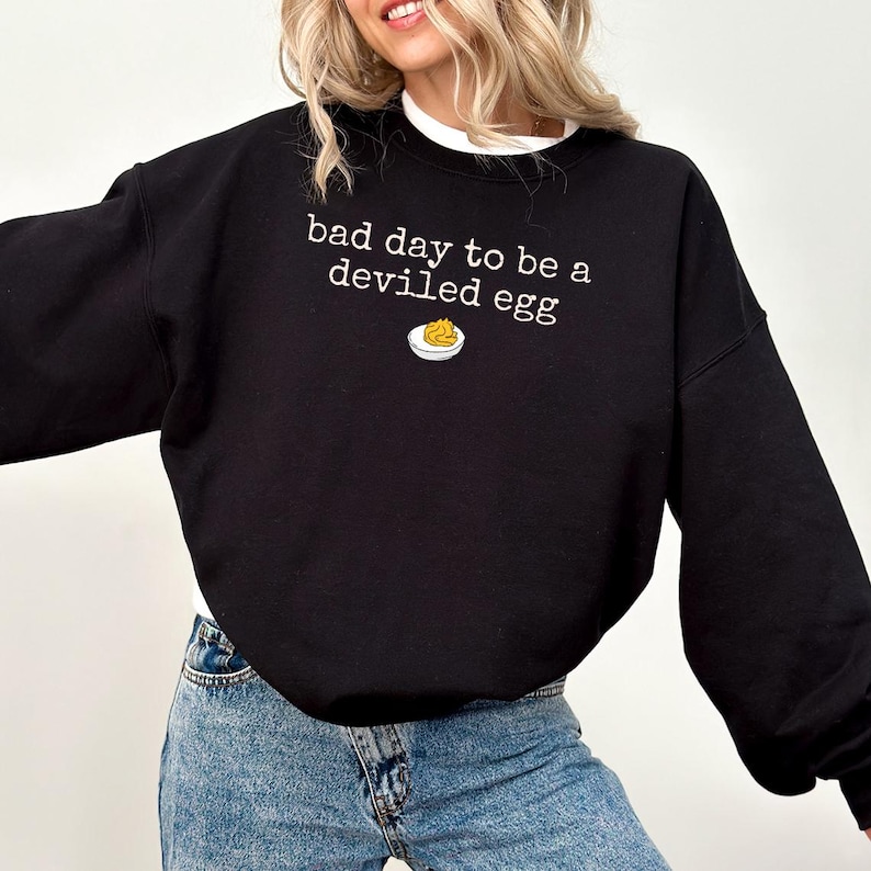 May include: Black sweatshirt with the phrase "bad day to be a deviled egg" in white text. A small graphic of a deviled egg is below the text. The sweatshirt is paired with blue jeans.