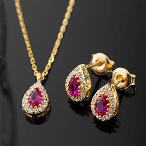 May include: A gold-colored jewelry set featuring a pendant necklace and matching earrings. Each piece has a teardrop-shaped, ruby-colored stone surrounded by small, clear stones. The set is displayed on a reflective black surface.