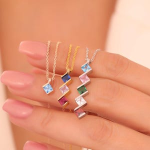 May include: A collection of gemstone pendant necklaces in various colors, including blue, pink, green, and white. The necklaces feature a geometric design with a small square gemstone pendant. The chains are gold and silver.