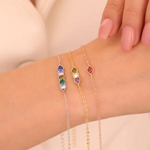 May include: Three delicate bracelets with colorful gemstones are displayed on a wrist. The bracelets are silver, gold, and rose gold, each featuring a different gemstone arrangement. The gemstones include blue, green, purple, and red, adding a vibrant touch to the jewelry.