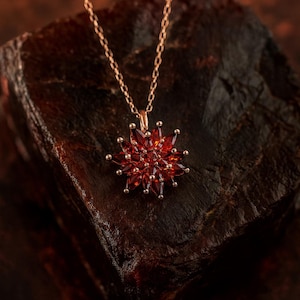 May include: A gold-toned necklace with a pendant shaped like a starburst. The pendant is made of numerous red gemstones. The necklace is displayed on a dark, textured surface.