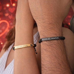 May include: Two matching bracelets are shown, one gold and one black. The gold bracelet is engraved with the date "04.03.2020". The black bracelet has Roman numerals. Both bracelets are on black cords.
