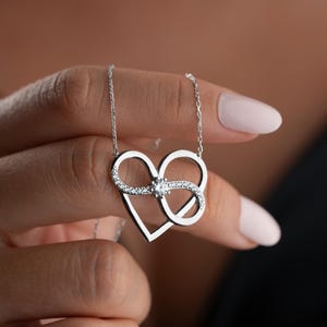 May include: A silver-toned necklace featuring a pendant shaped like a heart intertwined with an infinity symbol. The pendant is accented with small, clear, sparkling stones along the infinity loop and a single larger stone at the centre point.
