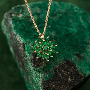 May include: A gold-colored necklace with a pendant shaped like a flower. The flower pendant is made of green gemstones and small gold-colored beads. The necklace is displayed on a rough, green stone.