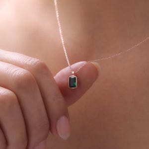 May include: A gold necklace with a rectangular emerald-cut green gemstone pendant. The pendant is set in a gold bezel and hangs from a fine gold chain. The necklace is shown against a blurred background.