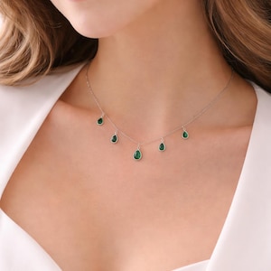 May include: A delicate silver necklace featuring six teardrop-shaped emerald green gemstones. The gemstones are evenly spaced along the chain, creating a subtle and elegant design. The necklace is worn on a person's neck.