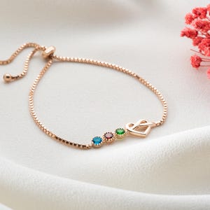 May include: Rose gold adjustable bracelet with a delicate chain. The bracelet features three colored gemstones: blue, red, and green, and a heart-shaped charm. The bracelet is displayed on a white, textured surface.