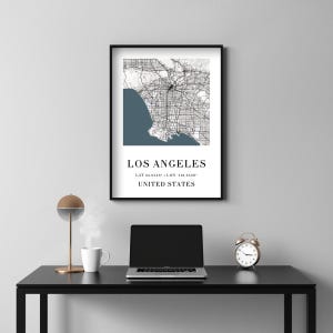 May include: A framed map print of Los Angeles, California, with the city's name and coordinates displayed below. The map features a black and white design with a blue coastal area. The print is in a black frame.