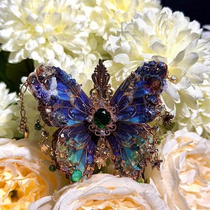 May include: A butterfly-shaped brooch featuring deep blue and teal translucent wings with intricate gold-toned metal filigree. A central oval emerald-green gemstone is surrounded by small clear crystals, with dangling charms and small green beads.
