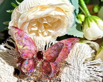 May include: A decorative butterfly brooch with pink and gold wings, a central red gem, and dangling charms. The brooch features intricate details and is embellished with rhinestones. The butterfly is set against a white, textured background.