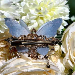 May include: A delicate butterfly hair clip with light blue wings and gold-colored metal accents. The clip features intricate detailing, including a decorative frame and dangling charms. The butterfly wings are translucent, adding a whimsical touch to this hair accessory.