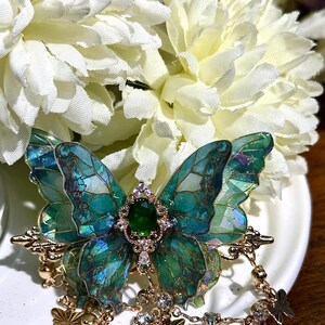 May include: A detailed, handcrafted butterfly brooch with iridescent teal and green wings. The brooch features a central emerald-colored gem and gold-toned accents, with dangling charms. The brooch is set against a backdrop of white flowers.