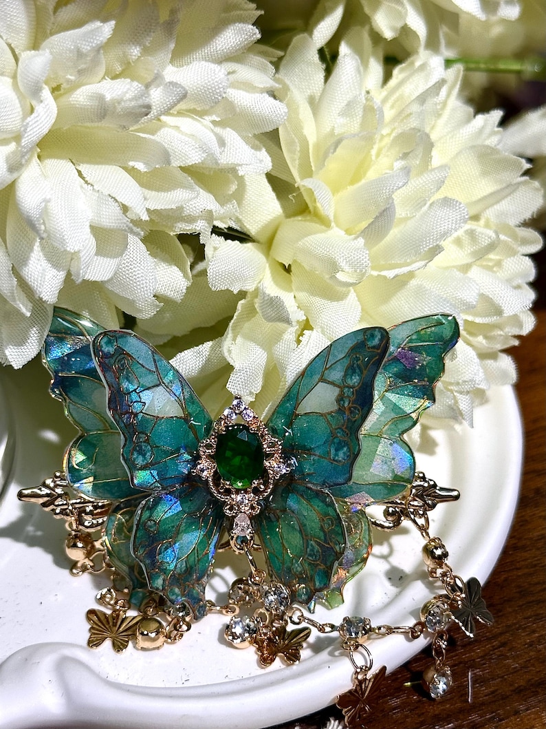 May include: A detailed, handcrafted butterfly brooch with iridescent teal and green wings. The brooch features a central emerald-colored gem and gold-toned accents, with dangling charms. The brooch is set against a backdrop of white flowers.