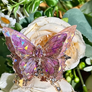May include: A decorative butterfly brooch with iridescent purple wings and gold-coloured accents. The brooch features a central gemstone and dangling crystal charms, set against a backdrop of white and cream-coloured flowers and greenery.