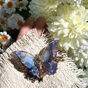 May include: A detailed butterfly brooch with iridescent blue and purple wings, accented with gold filigree and a central gemstone. The brooch is set on a cream-colored, fringed fabric, with white flowers in the background.
