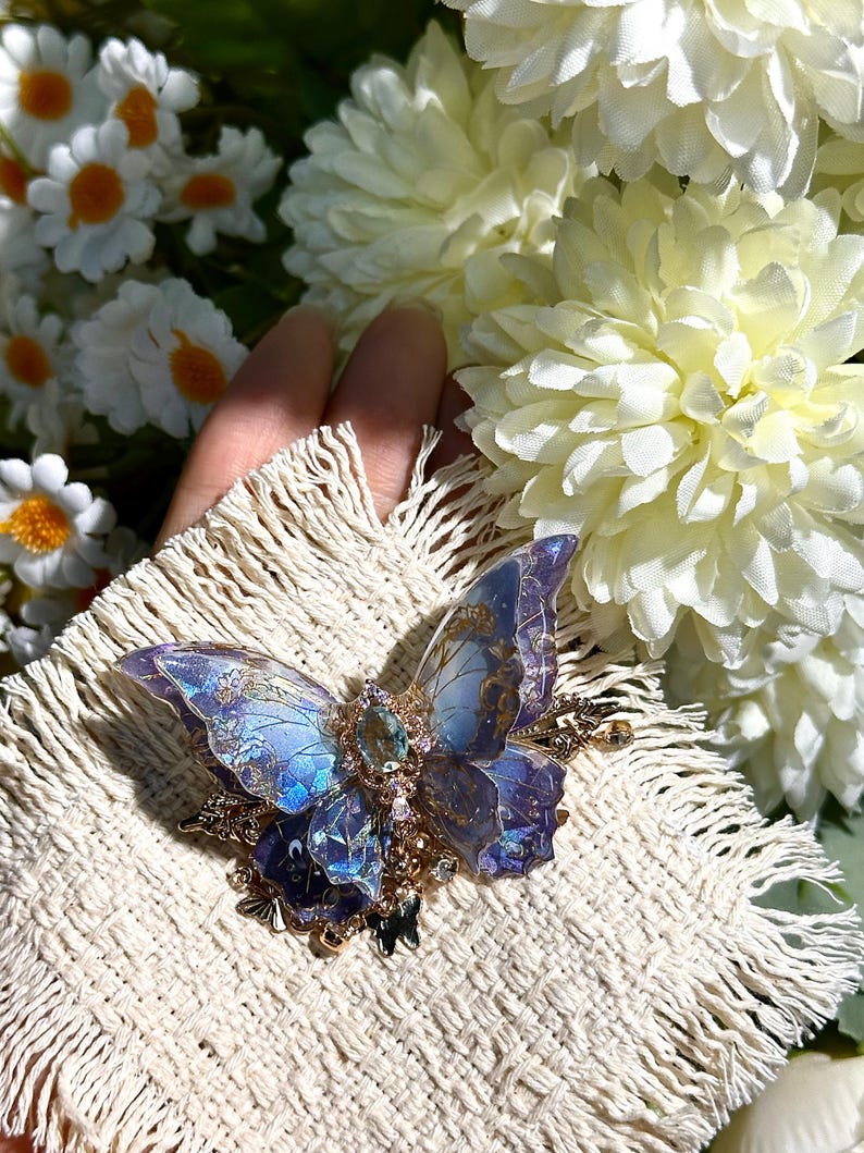 May include: A detailed butterfly brooch with iridescent blue and purple wings, accented with gold filigree and a central gemstone. The brooch is set on a cream-colored, fringed fabric, with white flowers in the background.