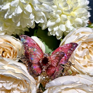 May include: A butterfly brooch with pink and purple iridescent wings, a central red gem, and gold filigree. The brooch is set against white and cream-coloured flowers.