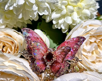 May include: A decorative butterfly brooch with iridescent pink and purple wings, a central red gem, and gold-colored filigree. The brooch is set against a backdrop of white and cream-colored flowers.