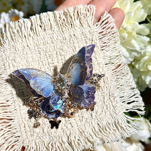 May include: A detailed butterfly brooch with blue and purple wings, accented with gold-colored metal and small dangling charms. The brooch is set on a cream-colored, fringed fabric, with a background of white and yellow flowers.