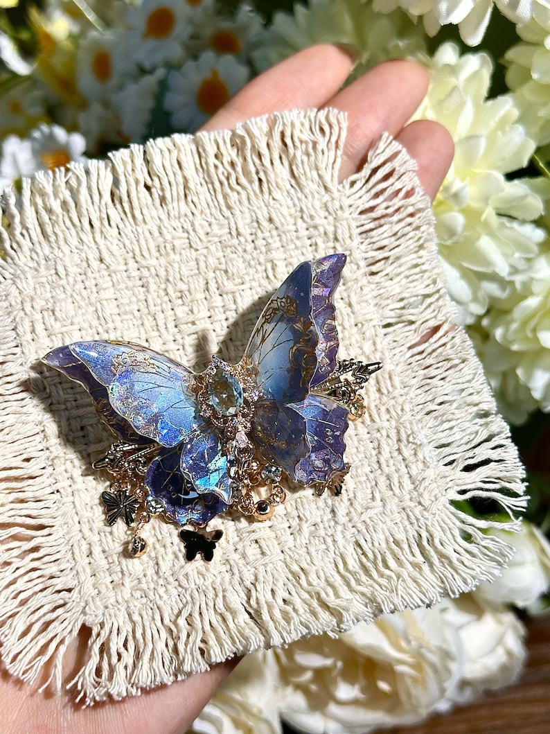 May include: A detailed butterfly brooch with blue and purple wings, accented with gold-colored metal and small dangling charms. The brooch is set on a cream-colored, fringed fabric, with a background of white and yellow flowers.