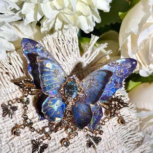 May include: A detailed butterfly brooch with iridescent blue wings and a central blue gemstone. The brooch features gold-colored accents, dangling chains with small butterflies, and clear crystal embellishments. The background includes white flowers.