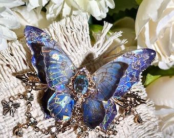 May include: A detailed butterfly brooch with iridescent blue wings and a central blue gemstone. The brooch features gold-colored accents, dangling chains with small butterflies, and clear crystal embellishments. The background includes white flowers.