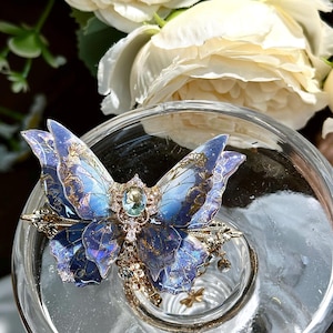 May include: A detailed butterfly-shaped ring with blue and gold accents, set with a central gemstone. The ring is placed inside a clear glass container, with white flowers in the background. The butterfly's wings have intricate patterns.