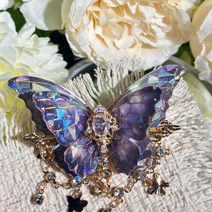 May include: A decorative butterfly brooch with iridescent purple wings and a gold-colored filigree body. The brooch features dangling clear crystals and small black butterfly charms, creating a whimsical and elegant accessory.