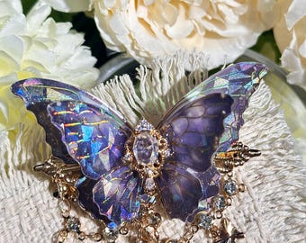 May include: A decorative butterfly brooch with iridescent purple wings and a gold-colored filigree body. The brooch features dangling clear crystals and small black butterfly charms, creating a whimsical and elegant accessory.
