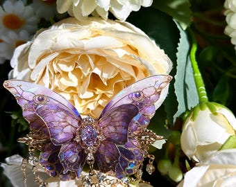 May include: A detailed, purple butterfly brooch with gold accents and dangling crystals. The butterfly has intricate wing patterns and a central jewel. The brooch is set against a backdrop of white and cream-colored flowers.