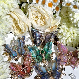 May include: A collection of colorful butterfly brooches with iridescent wings and gold-colored metalwork. The brooches are adorned with gemstones and are arranged on a white plate surrounded by white flowers.
