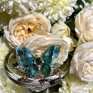 May include: A detailed butterfly brooch with iridescent teal wings, a central green gem, and gold-colored accents. The brooch is displayed on a clear glass pedestal, surrounded by white and cream-colored flowers.