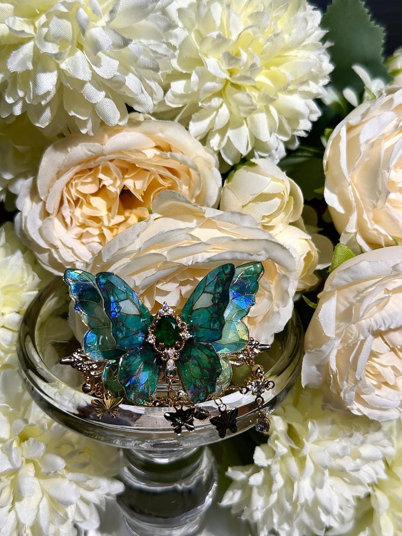 May include: A detailed butterfly brooch with iridescent teal wings, a central green gem, and gold-colored accents. The brooch is displayed on a clear glass pedestal, surrounded by white and cream-colored flowers.