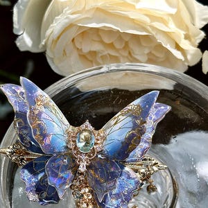 May include: A detailed close-up of a blue and gold butterfly-shaped hair clip with a central gemstone and intricate detailing. The clip is set against a blurred background with a white rose and a glass surface.