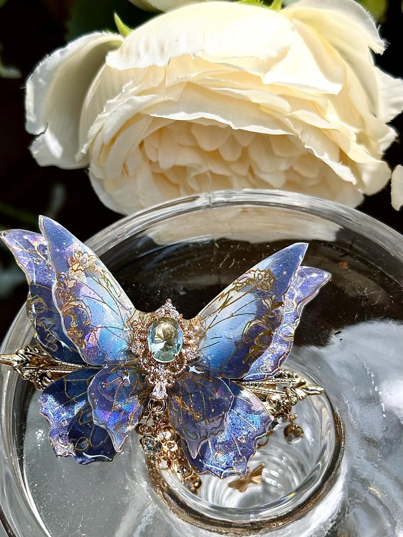 May include: A detailed close-up of a blue and gold butterfly-shaped hair clip with a central gemstone and intricate detailing. The clip is set against a blurred background with a white rose and a glass surface.