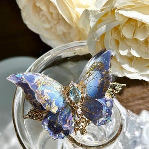 May include: A detailed, blue butterfly-shaped brooch with gold accents and a central gemstone. The wings feature intricate patterns and gradients of blue and purple. The brooch is set on a clear glass surface, with white flowers in the background.