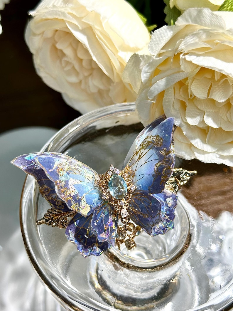 May include: A detailed, blue butterfly-shaped brooch with gold accents and a central gemstone. The wings feature intricate patterns and gradients of blue and purple. The brooch is set on a clear glass surface, with white flowers in the background.