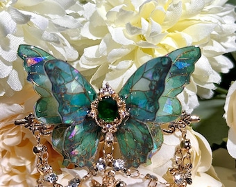 May include: A detailed butterfly brooch with teal and green wings, a central green gem, and gold-colored accents. The brooch features dangling charms with small butterflies and clear crystals. The background is a soft focus of white flowers.