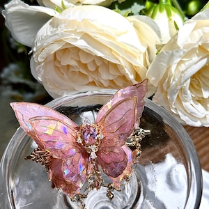 May include: A pink and gold butterfly hair clip with iridescent wings and jewel detailing. The butterfly sits on a clear glass surface, with white roses in the background. The clip has a delicate, ornate design.