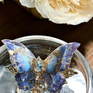 May include: A detailed butterfly hair clip with blue and purple wings, adorned with gold accents and a central jewel. The clip features intricate detailing and dangling charms, set against a blurred background.