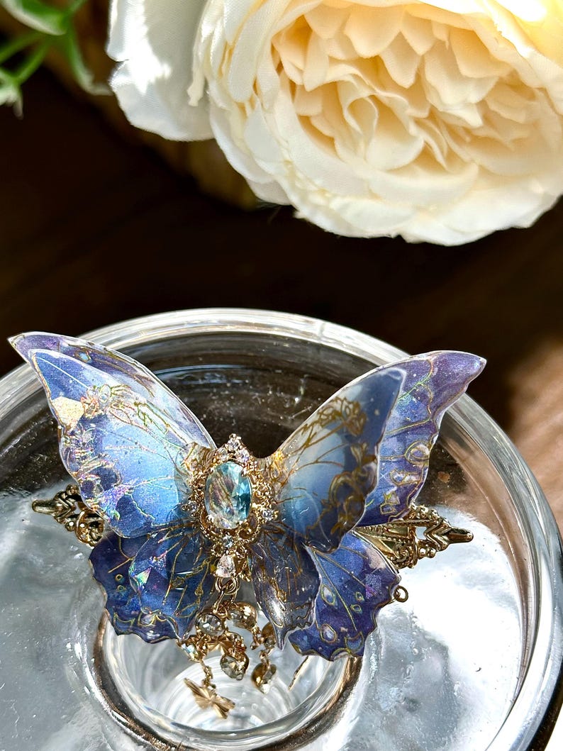 May include: A detailed butterfly hair clip with blue and purple wings, adorned with gold accents and a central jewel. The clip features intricate detailing and dangling charms, set against a blurred background.