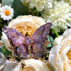 May include: A detailed butterfly brooch with purple and gold accents, adorned with sparkling crystals. The butterfly's wings feature intricate patterns and a central purple gemstone. The brooch is set against a backdrop of white and yellow flowers.
