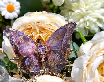 May include: A detailed butterfly brooch with purple and gold accents, adorned with sparkling crystals. The butterfly's wings feature intricate patterns and a central purple gemstone. The brooch is set against a backdrop of white and yellow flowers.