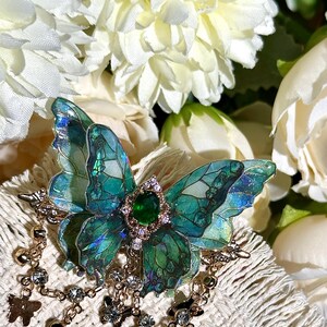 May include: A detailed butterfly brooch with iridescent teal and green wings, a central green gem, and a gold-colored frame. The brooch has dangling chains with small charms and crystals. The background features white flowers.