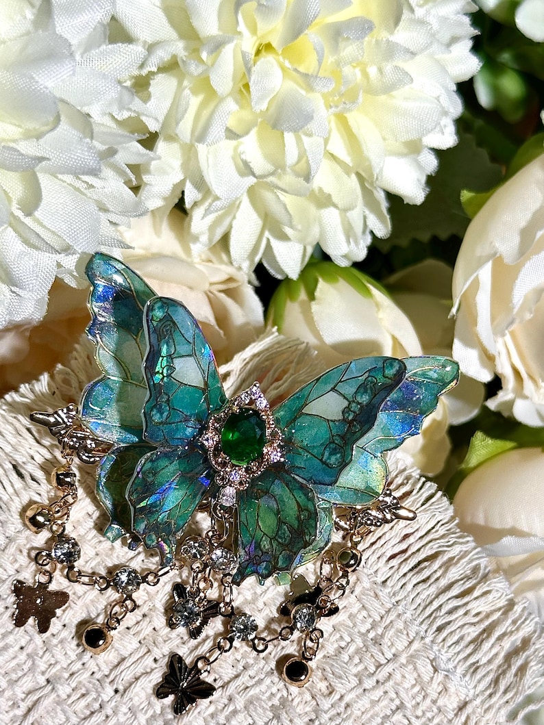 May include: A detailed butterfly brooch with iridescent teal and green wings, a central green gem, and a gold-colored frame. The brooch has dangling chains with small charms and crystals. The background features white flowers.
