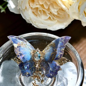 May include: A detailed butterfly-shaped accessory with blue and purple wings, adorned with gold filigree and a central blue gemstone. The butterfly has intricate patterns and is embellished with dangling charms. The accessory is set on a clear glass surface.