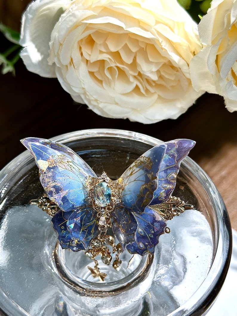 May include: A detailed butterfly-shaped accessory with blue and purple wings, adorned with gold filigree and a central blue gemstone. The butterfly has intricate patterns and is embellished with dangling charms. The accessory is set on a clear glass surface.